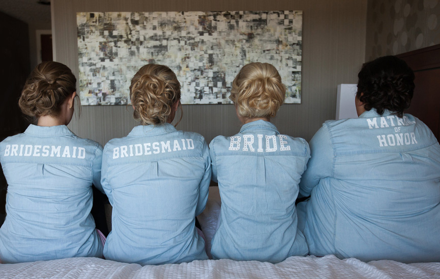 Bridal party in personalized chambray shirts sitting on a bed.